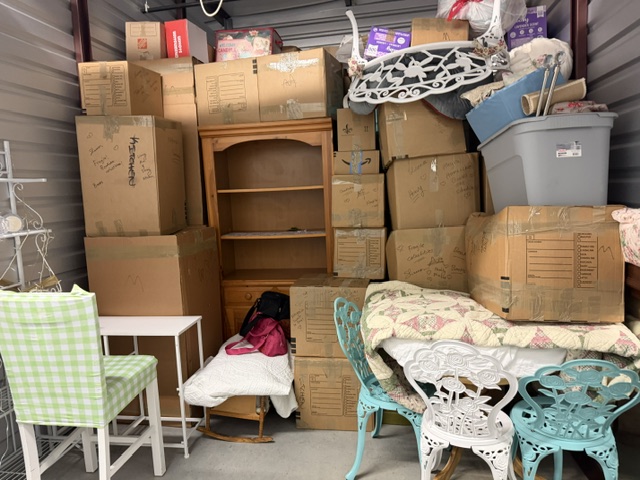 Crowded self-storage unit filled with cardboard boxes, wooden furniture, bedding, and household items