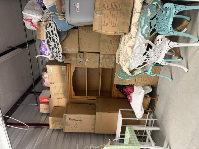 Cluttered storage garage filled with cardboard boxes, wooden furniture, and household items stacked on shelves