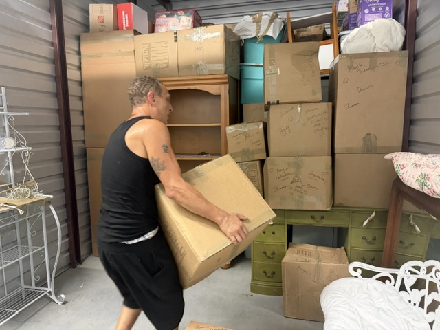 Man organizing cardboard boxes in a storage unit filled with furniture and household items