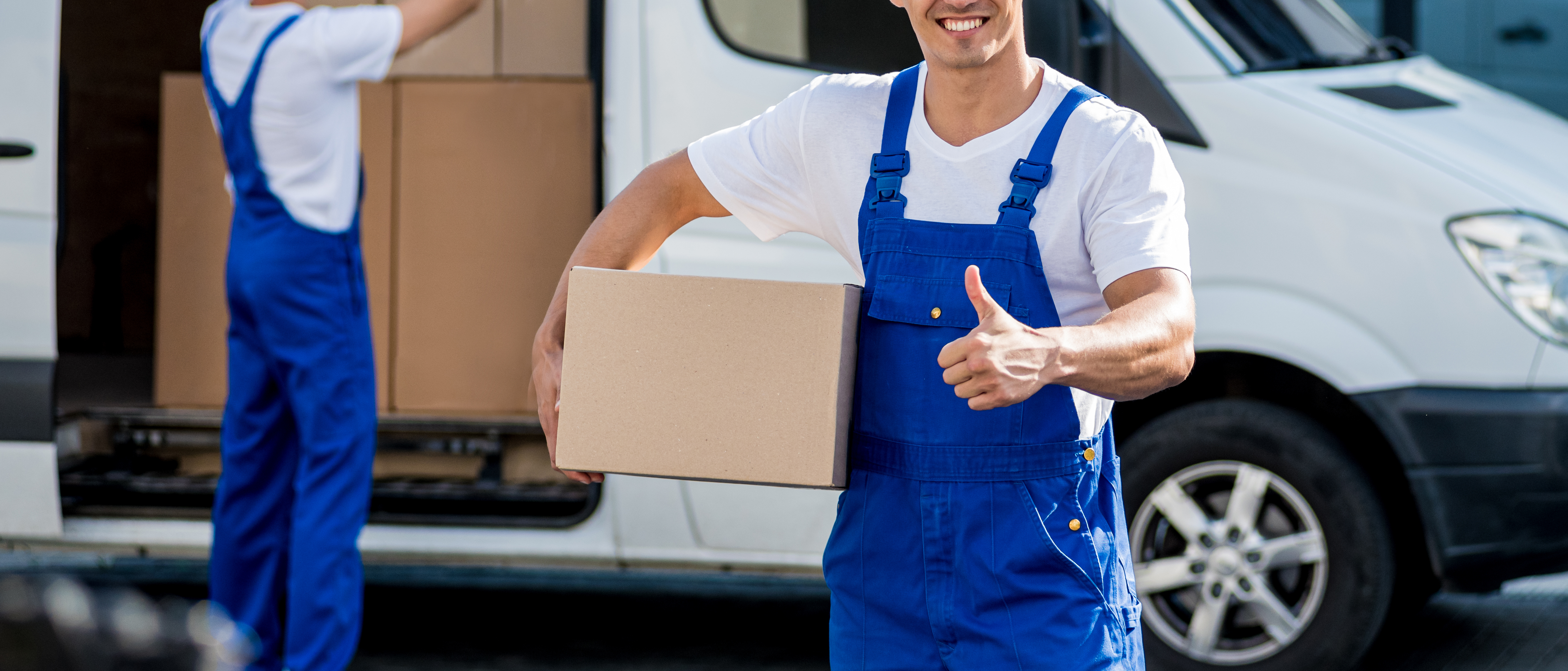 Smiling delivery worker in blue overalls holding cardboard box next to van