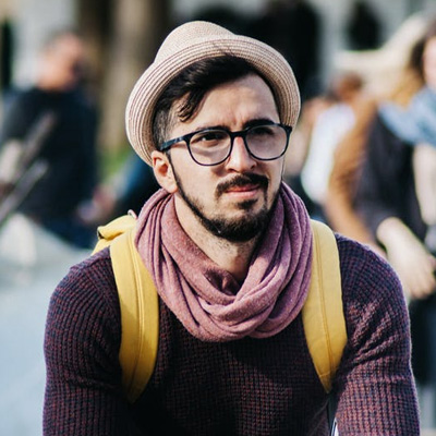 Man wearing glasses, beige hat, purple scarf, and yellow backpack on urban street